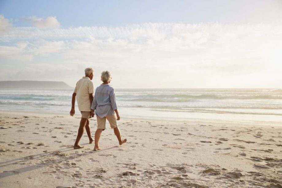 Rear View Of Senior Couple Walking Along Beach Hand In Hand